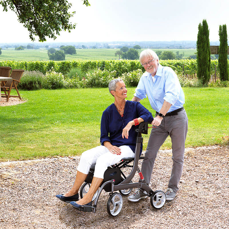 Modell Spring mit Variopaket von Bescomedical, Front-/ Seitenansicht eines grauen Rollators in einem Park, mit sitzender Person und schiebender Person